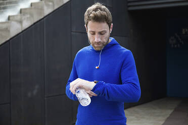 Portrait of jogger with water bottle, checking workout progress on his smart watch - JSRF00652
