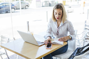 Junge Frau mit Laptop und Notizbuch auf einem Tisch in einem Cafe - LJF01049