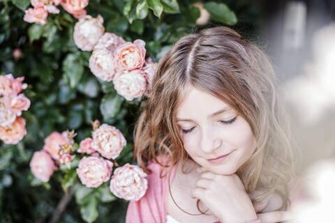 Porträt eines Mädchens mit geschlossenen Augen neben einem rosa Rosenstrauch, lizenzfreies Stockfoto