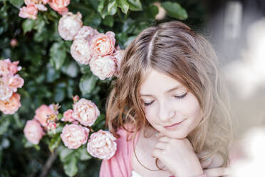 Portrait of girl with eyes closed beside pink rosebush - STBF00419