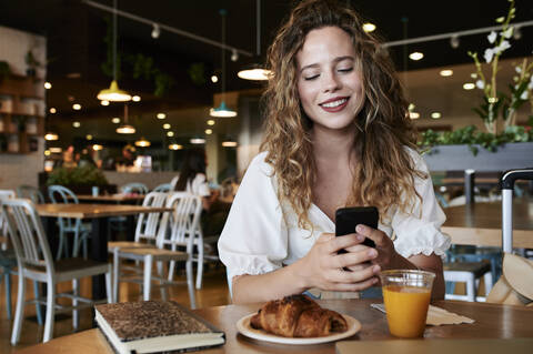 Lächelnde junge Frau benutzt ihr Smartphone in einem Café beim Frühstück, lizenzfreies Stockfoto