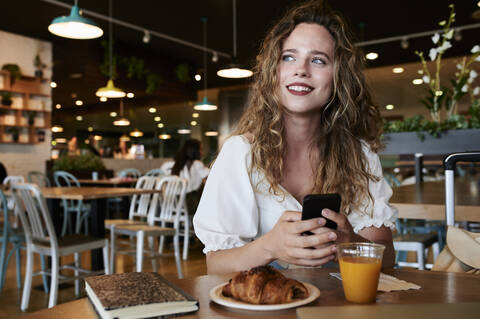 Lächelnde junge Frau mit Smartphone in einem Cafe beim Frühstück, lizenzfreies Stockfoto