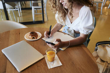 Junge Frau in einem Café, die ihr Smartphone benutzt und sich Notizen macht, während sie frühstückt - IGGF01335