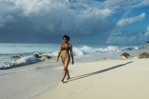 Frau im Bikini spaziert am Strand von Grace Bay gegen bewölkten Himmel, Providenciales, Turks- und Caicosinseln - RUNF03344