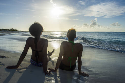 Rückansicht von Freundinnen, die bei Sonnenuntergang am Strand von Grace Bay auf das Meer blicken, Providenciales, Turks- und Caicosinseln - RUNF03346