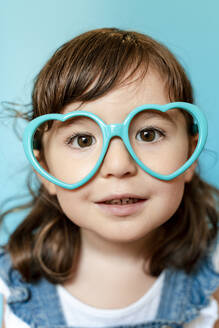 Portrait of cute little girl with heart shaped glasses on blue background - GEMF03176