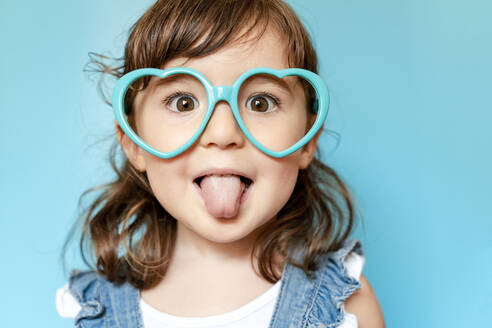 Portrait of cute little girl with tongue out and heart shaped glasses on blue background - GEMF03178