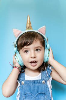 Portrait of cute little girl listening music and singing with unicorn shaped earphones on blue background - GEMF03180