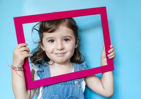 Portrait of cute little girl holding a picture frame on blue background - GEMF03181