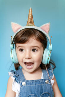 Portrait of cute little girl listening music and singing with unicorn shaped earphones on blue background - GEMF03184