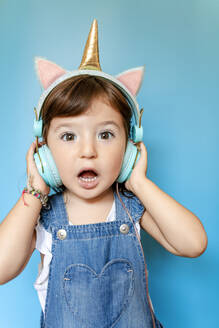 Portrait of cute little girl listening music and singing with unicorn shaped earphones on blue background - GEMF03185