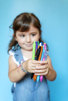 Portrait of cute little girl picking up a handful of colored ballpoint pens on blue background - GEMF03186