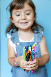 Porträt eines süßen kleinen Mädchens, das eine Handvoll bunter Kugelschreiber auf blauem Hintergrund aufhebt - GEMF03187