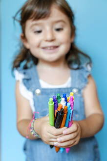 Portrait of cute little girl picking up a handful of colored ballpoint pens on blue background - GEMF03187