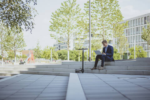 Young businessman with e-scooter using laptop in the city stock photo