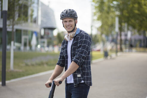 Porträt eines lächelnden jungen Mannes mit E-Roller in der Stadt - MOEF02463