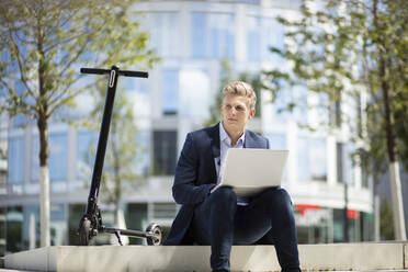 Young businessman with e-scooter using laptop in the city - MOEF02491