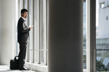 Junger Geschäftsmann mit Mobiltelefon am Fenster stehend - JPIF00199