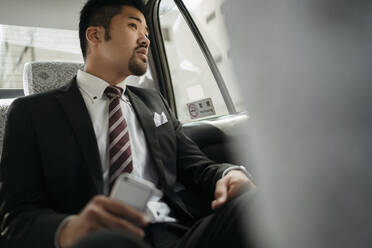 Junger Geschäftsmann mit Mobiltelefon in einem Taxi, der aus dem Fenster schaut - JPIF00214