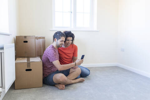 Glückliches Paar sitzt auf dem Boden im neuen Haus und benutzt ein Handy, lizenzfreies Stockfoto