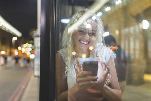 Junge Frau in London bei Nacht, die auf ihr Smartphone schaut und auf den Bus wartet - WPEF01988