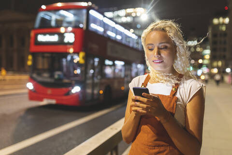 Junge Frau in London bei Nacht, die auf ihr Smartphone schaut, lizenzfreies Stockfoto