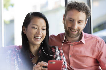 Happy man and woman with cell phone and earbuds in a cafe - FKF03653