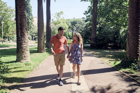 Lächelndes junges Paar geht Hand in Hand durch einen Park - JNDF00125
