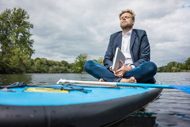 Geschäftsmann mit Laptop auf SUP-Board auf einem See sitzend - JOSF03782