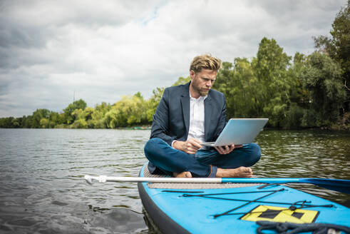 Geschäftsmann sitzt auf einem SUP-Board auf einem See und benutzt einen Laptop - JOSF03790