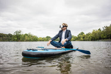 Geschäftsmann mit VR-Brille sitzt auf einem SUP-Board auf einem See - JOSF03795