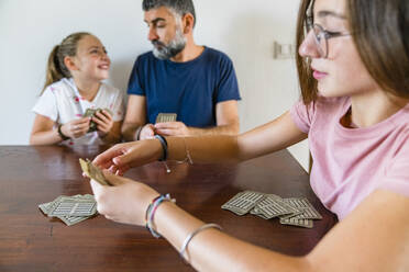 Vater mit zwei Töchtern spielt Karten auf einem Holztisch zu Hause - MGIF00701