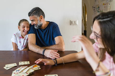 Glücklicher Vater mit zwei Töchtern spielt Karten auf einem Holztisch zu Hause, lizenzfreies Stockfoto