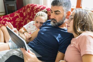 Vater mit zwei Töchtern, die zu Hause auf der Couch auf ein Tablet schauen - MGIF00706