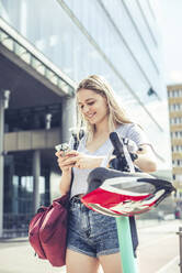 Porträt einer lächelnden jungen Frau, die sich auf den Lenker eines E-Scooters stützt und dabei ein Mobiltelefon benutzt, Berlin, Deutschland - BFRF02073