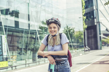 Porträt einer glücklichen jungen Frau mit Fahrradhelm, die sich auf den Lenker eines E-Scooters stützt - BFRF02076