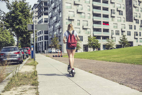 Rückenansicht einer jungen Frau mit Rucksack auf einem E-Scooter auf dem Bürgersteig, Berlin, Deutschland - BFRF02077