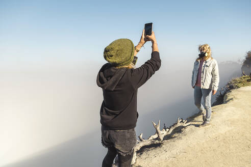 Junge Frau mit Atemschutzmaske beim Fotografieren ihrer Freundin am Rande des Vulkans Ijen, Java, Indonesien - KNTF03600