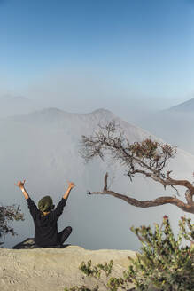 Frau mit Atemschutzmaske sitzt am Vulkan Ijen und hebt die Arme, Java, Indonesien - KNTF03613