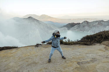 Junge mit Atemschutzmaske steht mit ausgestreckten Armen am Rand des Vulkans Ijen, Java, Indonesien - KNTF03619
