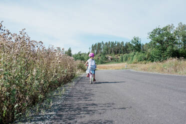 Rückansicht eines jungen Mädchens, das auf der Landstraße von der Schule nach Hause radelt - CAVF63653