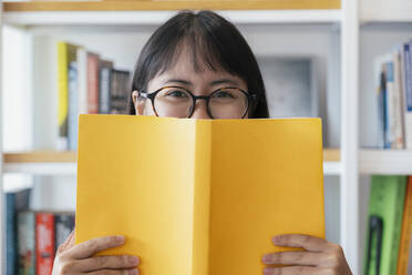 Fröhliche junge Frau bedeckt ihr Gesicht mit einem gelben Lehrbuch. - CAVF63723