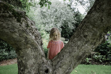 Young girl looking sad stood behind a tree in the rain in summer - CAVF63868