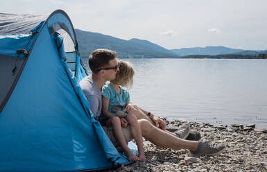 Ein Vater, der seine Tochter umarmt, während er in einem Zelt auf dem Campingplatz am Meer sitzt - CAVF63876