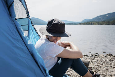 Frau in einem Zelt sitzend und lächelnd mit Sonnenhut beim Camping - CAVF63885