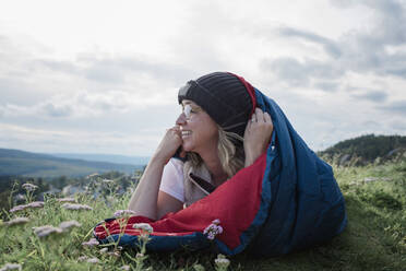 Frau liegt im Schlafsack auf einem Berg und schaut lächelnd auf die Aussicht - CAVF63889