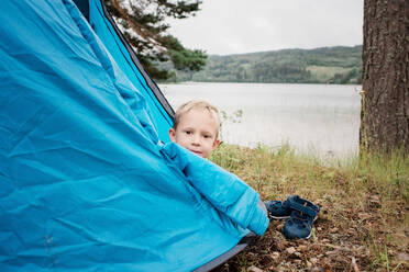 Porträt eines jungen blonden Jungen, der in einem Zelt am Wasser zeltet - CAVF63912