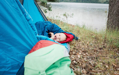 Porträt eines kleinen Jungen im Schlafsack beim Zelten am Wasser - CAVF63913