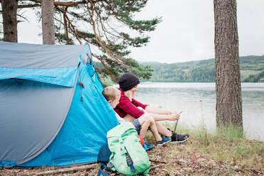 Mutter und Sohn sitzen in einem Zelt und binden lächelnd die Schnürsenkel beim Camping - CAVF63914