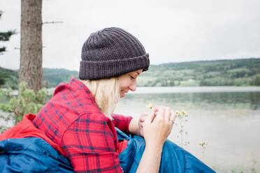 Frau betrachtet lächelnd eine Blume, während sie in einem Schlafsack beim Camping sitzt - CAVF63920